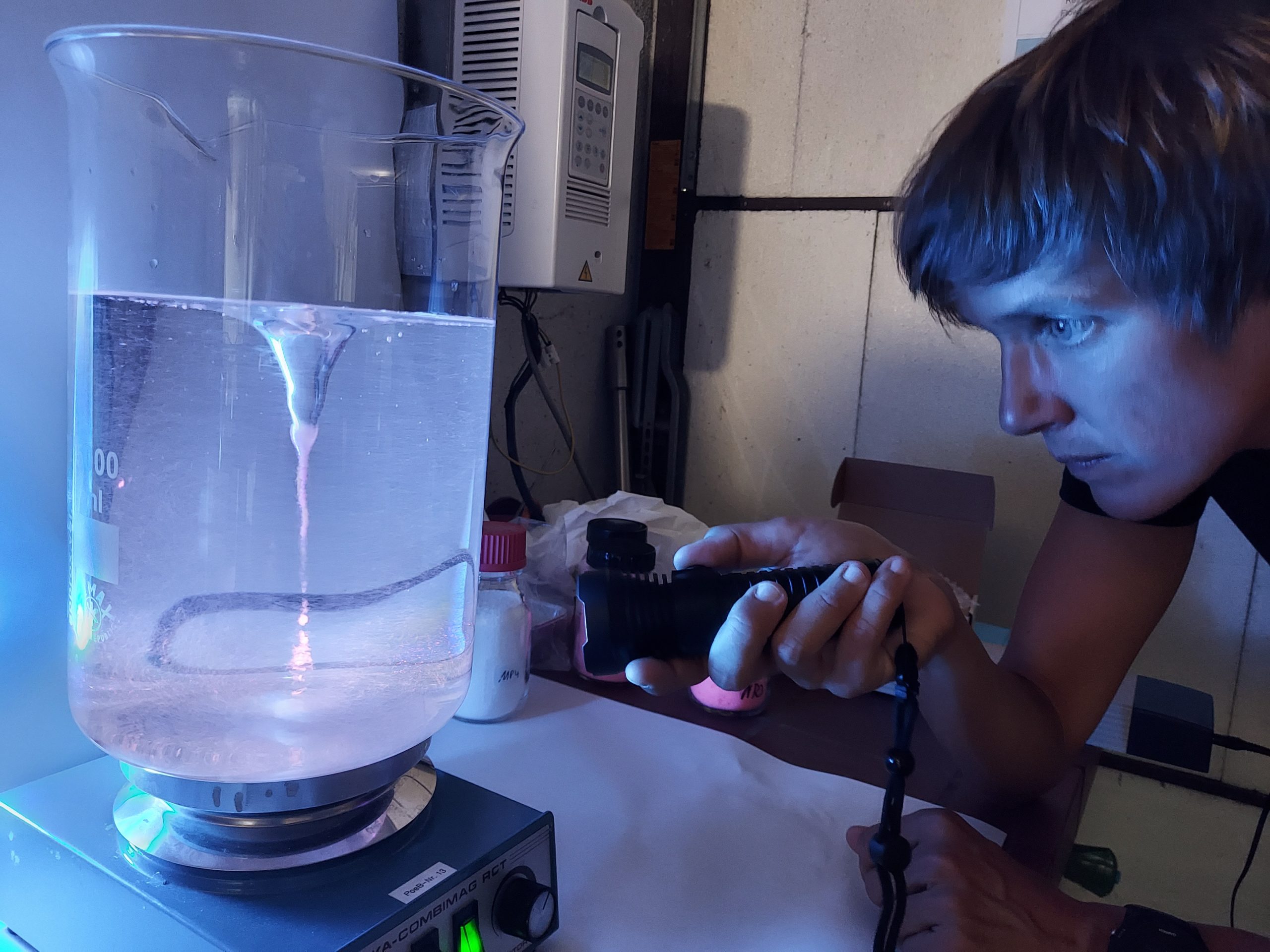 Katrin Schuhen im Labor vor einem 2l Wasserbehälter, in dem sie einen Strudel mit einer Taschenlampe beleuchtet und dadurch angefärbtes Mikroplastik erkennbar wird